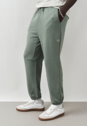 Mężczyzna ubrany w joggery koloru szałwii, białe skarpetki, białe sneakersy z gumowymi podeszwami oraz srebrną bransoletkę z łańcuszkiem, stojący z rękami w kieszeniach.