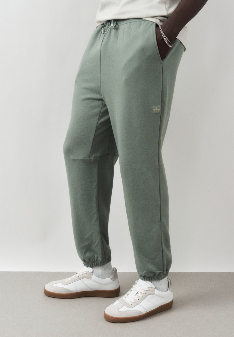 Άνδρας που φοράει χακί (φασκόμηλου) παντελόνι jogger, λευκές κάλτσες, λευκά αθλητικά παπούτσια με λαστιχένιες σόλες και ασημένιο βραχιόλι αλυσίδα, στέκεται με τα χέρια στις τσέπες.