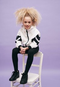 Bela zip-up jakna s crnim prugama, crne tajice i crne tenisice. Kovrčava plava kosa, sjedi na bijeloj stolici s ljubičastom pozadinom.