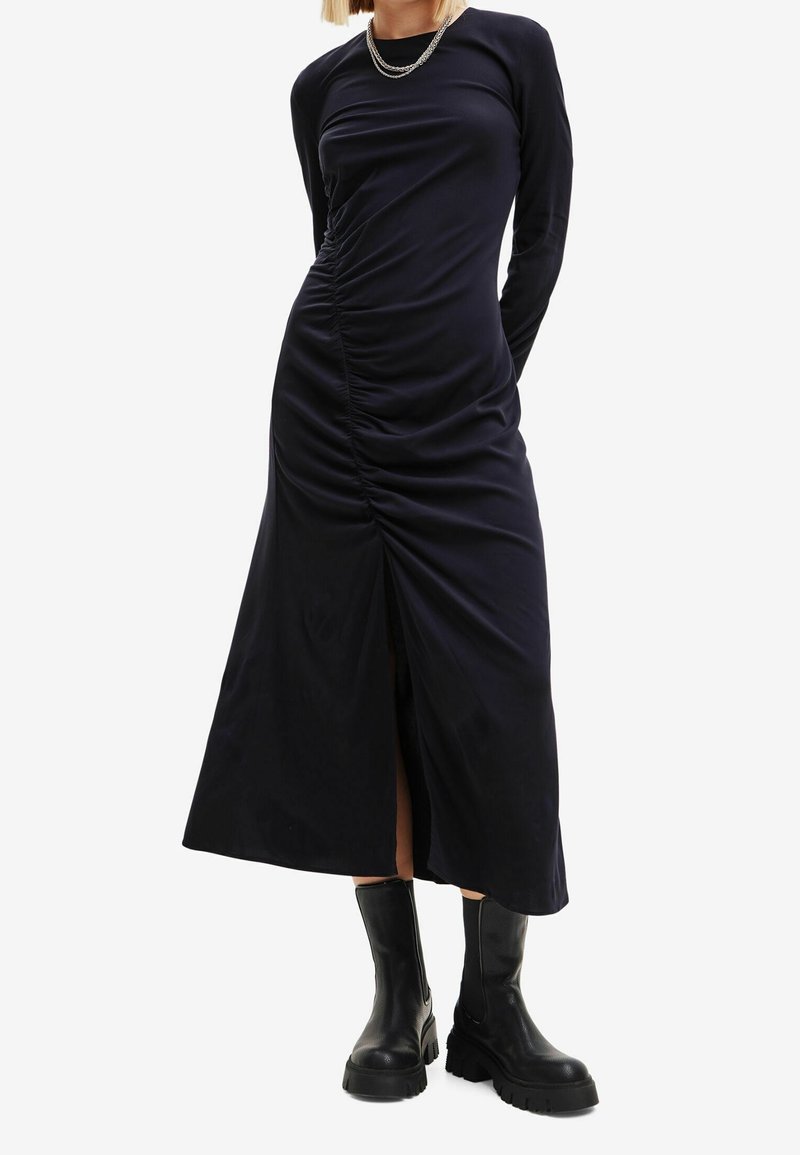 Femme portant une longue robe noire froncée avec une fente à l'avant et des bottes en cuir chunky noires, se tenant avec les mains derrière le dos.