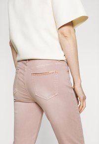 Jeans de color rosa pálido con una forma ajustada, que presentan bolsillos traseros decorados con un patrón metálico similar a una cadena y detalles de costura.
