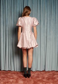 Rosa satengkjole med puffede ermer, utsvingt skjørt og en glatt finish, kombinert med mørkegrønne sokker og klumpete svarte hæler mot blå bakgrunn.