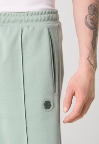 Pantalon de sport vert clair avec un tissu texturé, poche zippée sur le côté et un patch logo circulaire sur le bas de la jambe. Ceinture élastique visible.