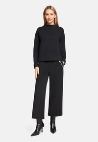 Sweat-shirt noir à col montant et épaules tombantes, associé à un pantalon noir à jambes larges. Tissu texturé avec un détail de logo subtil.