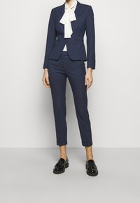 Granatowy garnitur na miarę z dopasowaną marynarką, wąskimi spodniami podwiniętymi przy mankietach oraz białą apaszką zawiązaną na szyi. Czarne buty z lakierowanej skóry.