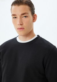 Czarny materiałowy sweatshirt z okrągłym dekoltem, z białą wewnętrzną naszywką. Tkanina wydaje się gruba i trwała. Model z neutralnym wyrazem twarzy.