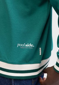 Groene sweatshirt met gestreepte witte en groene onderkant. Bevat het 'poolside.' logo in wit en roze, met een label waarop "REDEFINED REBEL" staat.
