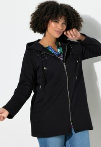 Giacca nera con zip e cappuccio, con dettagli in argento, coulisse e inserti intrecciati sulle spalle. Indossata sopra una camicia colorata a motivo.