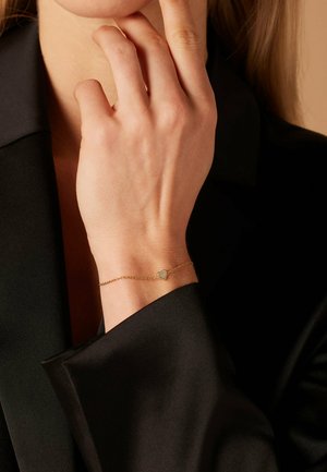 Bracelet en or avec une chaîne délicate et un petit pendentif en forme de cœur. Porté sur une main sur un tissu satin noir, mettant en valeur les détails fins.