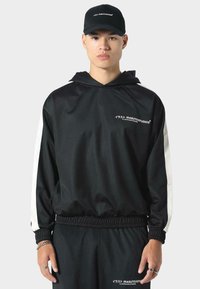 Giacca nera con cappuccio, strisce laterali bianche e polsini elasticizzati, con logo sul petto. Abbinata a pantaloni della tuta neri e un berretto coordinato.