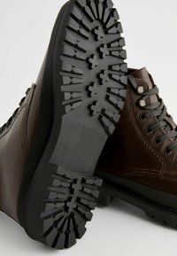 Botas de cuero marrón con una suela resistente, una textura de goma negra en la suela, y cordones grises. Presenta ojetes metálicos redondos y un diseño de tobillo alto.