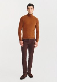Brązowy sweter z golfem, dopasowany krój, zestawiony z ciemnobrązowymi spodniami o slim-fit. Model stoi na neutralnym tle, eksponując proste akcenty.