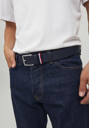 Marinblå belte med en sølvmetallspenne, med et rødt, hvitt og blått stripete akksent. Båret over mørke denim jeans.