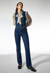 Chaleco de mezclilla y jeans acampanados en azul oscuro, combinados con una blusa de color claro con un patrón que presenta un gran lazo. Acabados texturizados presentes.