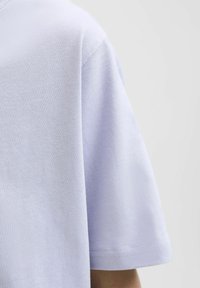 Šviesiai violetinė trumpomis rankovėmis marškinėlė, pagaminta iš minkštos medvilnės medžiagos, su apvalia iškirpte ir pastebimu siuvimu palei apykaklę ir rankoves.