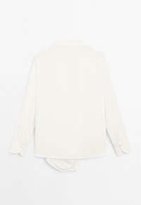 Blusa bianca a maniche lunghe con polsini abbottonati, colletto classico e orlo asimmetrico appesa su uno sfondo bianco.