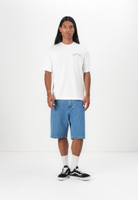 Hvid bomulds t-shirt med et lille trykt logo, parret med blå denimshorts. Modellen har sorte sneakers og hvide ankelsokker på.