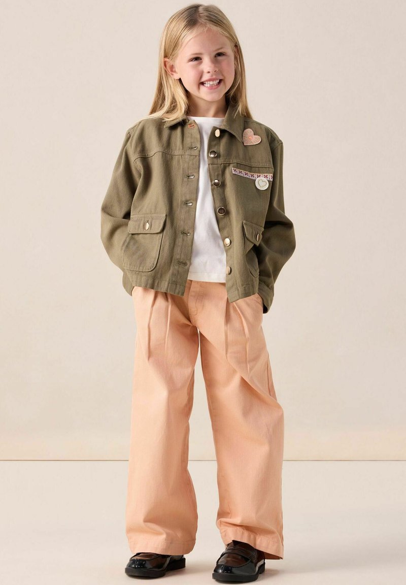 Veste vert olive avec boutons sur le devant, deux poches et patches décoratifs. Pantalons larges pêche clair et chaussures noires brillantes complètent la tenue.