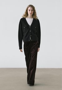 Zwarte gebreide cardigan met gouden knopen en twee voorzakken, gecombineerd met donkerbruine wijde gebreide broek van ribfluweel. Model staat tegen een minimalistische achtergrond.