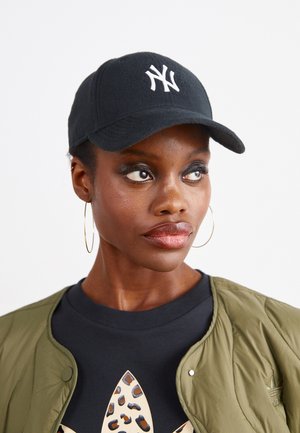 Casquette noire avec un logo "NY" blanc, en tissu texturé, avec une visière courbée, portée avec une veste bomber verte et un haut noir présentant un motif léopard.