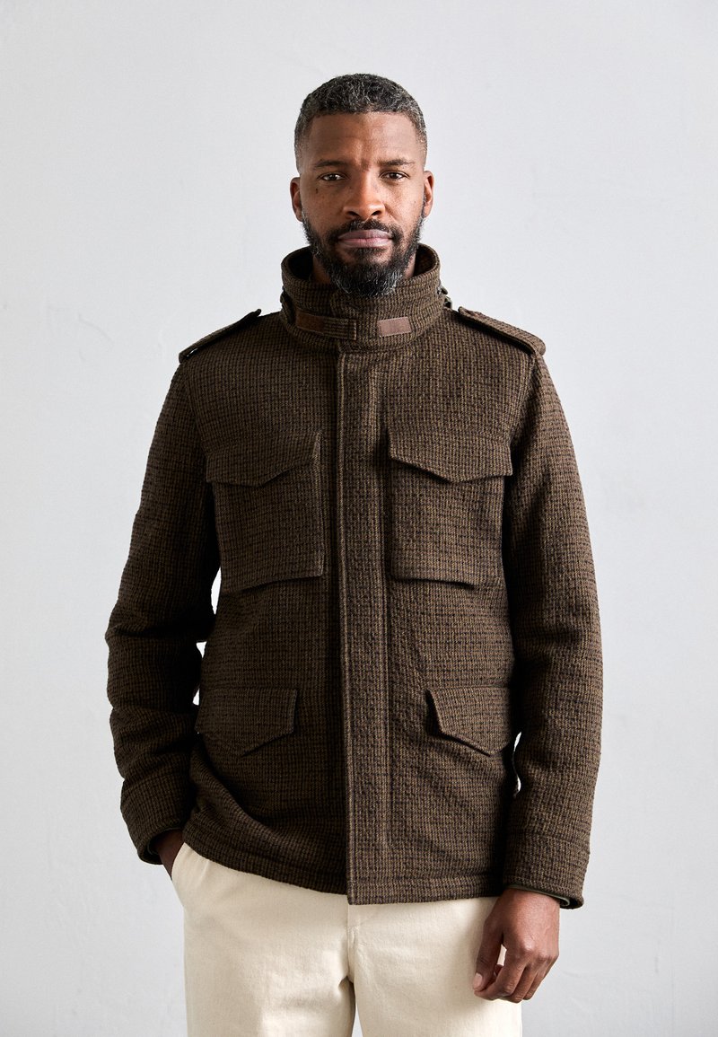 Veste texturée marron avec deux poches avant, col montant et épaulettes. Portée sur un pantalon crème, sur un fond uni.