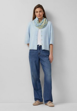 Woman standing wearing light blue buttoned cardigan, white top, floral scarf, blue jeans, and beige shoes against gray wall.
