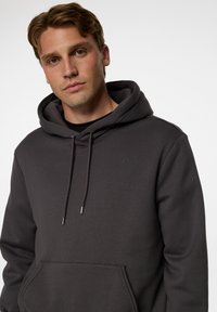 Mörkgrå huvtröja i mjukt tyg; har en framficka i kangurostil, dragsko på huvan och en subtil logotyp i matchande färg.