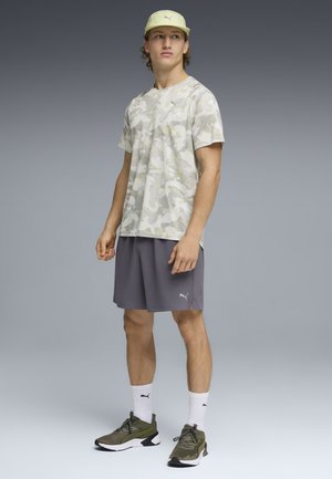 Bărbat tânăr purtând un tricou deschis la culoare cu model camuflaj, pantaloni scurți gri, șosete albe, adidași verde-olive și o șapcă bej, stând în picioare pe un fundal gri.