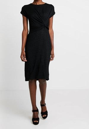 Kobieta w czarnej sukience do kolan z detalem skrętu z przodu, w połączeniu z czarnymi sandałami na obcasie z odkrytymi palcami i paskami wokół kostek.
