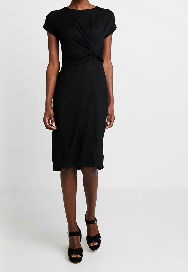 Femme portant une robe noire longueur genoux avec un détail torsadé à l'avant, associée à des sandales à talons ouvertes noires avec des brides à la cheville.