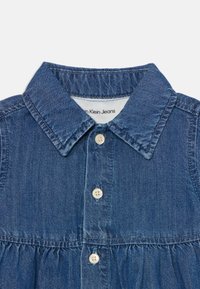 Denim košulja plave nijanse, s ovratnikom, prednjim kopčanjem na četiri kremaste gumbe i detaljem skupljenog struka.