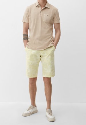 Polo beige avec patte à trois boutons et poche, associé à un short jaune clair avec un motif discret de feuilles. Baskets blanches.