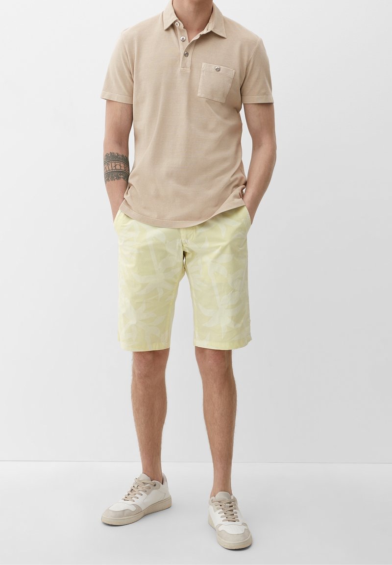 Polo beige avec patte à trois boutons et poche, associé à un short jaune clair avec un motif discret de feuilles. Baskets blanches.