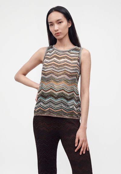 Jeune femme aux longs cheveux foncés portant un haut sans manches à motif zigzag multicolore et un pantalon sombre à motif zigzag, debout devant un fond blanc uni.