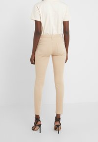 Jeans beige ajustados con cintura alta, bolsillos traseros y una textura suave, combinados con tacones altos negros de tiras y una camiseta de color claro.