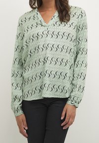 Vaaleanvihreä pusero mustalla aaltoilevalla kuviolla, V-pääntiellä ja pitkillä hihoilla, valmistettu sileästä kankaasta, yhdistetty tummiin farkkuihin.