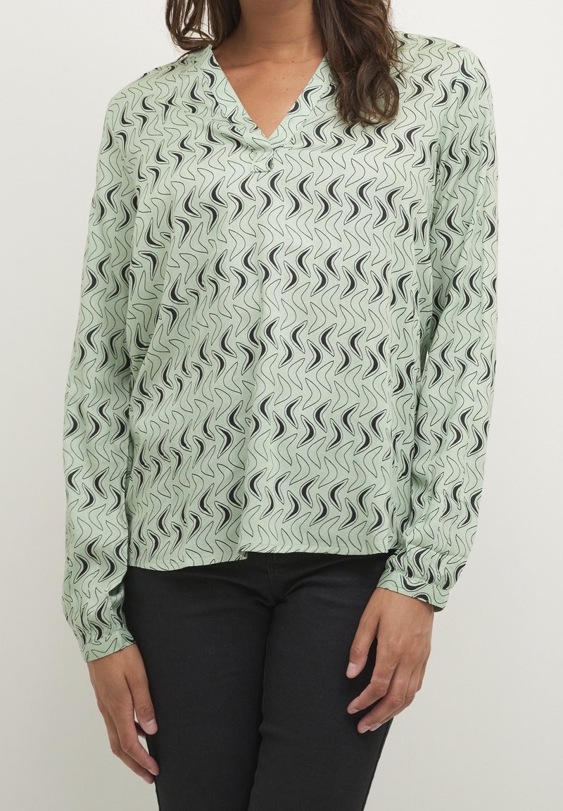 Vaaleanvihreä pusero mustalla aaltoilevalla kuviolla, V-pääntiellä ja pitkillä hihoilla, valmistettu sileästä kankaasta, yhdistetty tummiin farkkuihin.