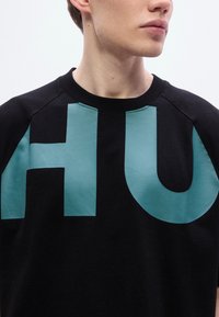 Μαύρο φούτερ με μεγάλο μπλε-πράσινο τύπωμα "HU" στο μπροστινό μέρος. Λαιμόκοψη τύπου crew με μανίκια raglan. Απαλή υφή υφάσματος.