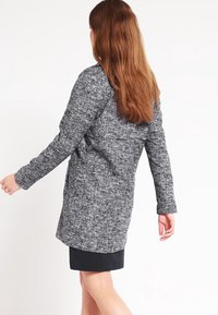 Cappotto grigio testurizzato con taglio dritto, maniche lunghe e tasche laterali, indossato sopra una gonna nera, visto da dietro.