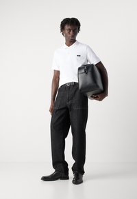Weißes Polohemd, schwarze Denim-Jeans, schwarze Lederschuhe und eine strukturierte schwarze Tasche. Bemerkenswerte Nähte an den Jeans und ein poliertes Taschendesign.