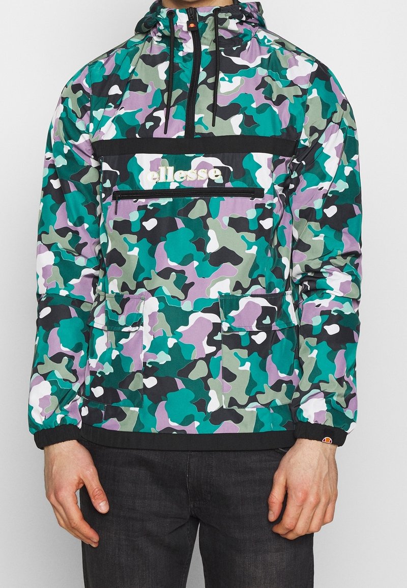 Homme portant une veste à capuche camouflage verte, violette, noire et blanche avec des poches avant zippées et le logo ellesse sur la poitrine.