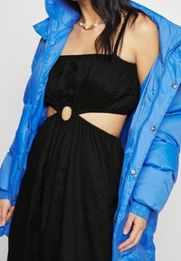 Modrá péřová bunda přes černé šaty s výřezem, které mají kruhový detail v oblasti pasu. Tkanina je hladká s mírným leskem.