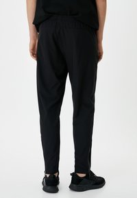 Schwarze Sporthose mit elastischem Bund, schmaler Passform und Knöchelabschlüssen. Das Material wirkt glatt und leicht, kombiniert mit schwarzen Turnschuhen.
