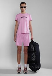 Conjunto de camiseta de algodón rosa y pantalones cortos con logo. El modelo sostiene un bolso negro con un estampado gráfico. Zapatillas multicolores y calcetines deportivos completan el look.