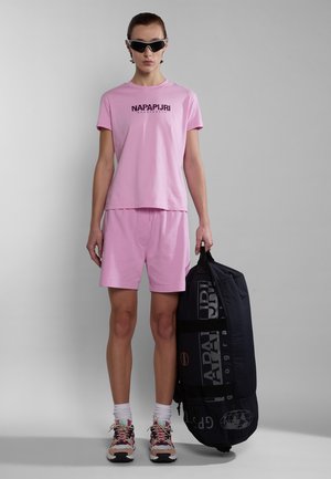 Rosa Baumwoll-T-Shirt und Shorts-Set mit Logo. Das Modell hält eine schwarze Tasche mit grafischem Druck. Bunte Sneakers und Sportsocken runden das Outfit ab.