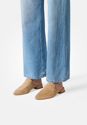 Person iført lyseblå vidde-ben jeans og brune ruskind backless loafers, der står på et hvidt gulv.