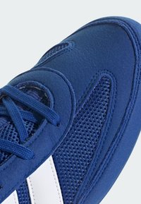 Gros plan sur l'avant d'une basket bleue avec tissu en maille, superpositions en daim et bandes blanches sous des lacets bleus sur un fond clair.