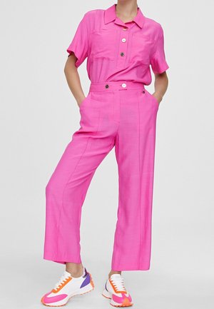 Person iført lyserød kortærmet skjorte med knapper og matchende bukser samt hvide, orange og lilla sneakers, stående med hænderne i lommerne.