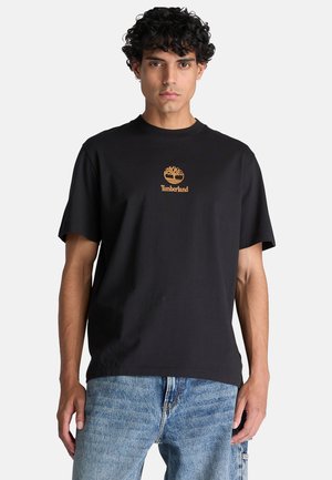 Ung mand med krøllet hår iført en sort Timberland t-shirt og blå jeans, står mod en ensfarvet lys baggrund.