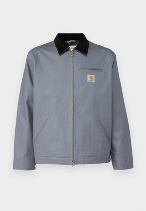 Chaqueta de algodón gris con un cuello de pana negro, cremallera frontal y bolsillo en el pecho con un logo amarillo de Carhartt. Incluye dos bolsillos laterales.